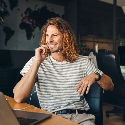Young curly man using laptop and earphones while sitting by desk in office
